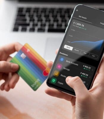 Eine Hand hält eine bunte bunq-Karte, während die andere ein Smartphone mit einer Finanzübersicht zeigt. Die App zeigt ein Guthaben von 1.559,76 € sowie eine grafische Darstellung der Vermögensentwicklung. Im Hintergrund ist ein Laptop sichtbar.