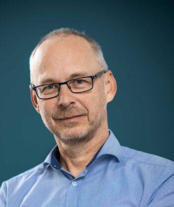 Ein Mann mit Brille und einem blauen Hemd blickt in die Kamera. Der Hintergrund ist einfarbig und blau. Die Darstellung vermittelt einen professionellen Eindruck, der im Kontext der Diskussion über adaptive Angriffssysteme relevant ist.