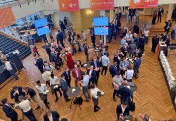 Das Point Zero Forum versammelt zahlreiche internationale Teilnehmer in einem Kongresshaus. Die Atmosphäre ist geprägt von Networking und Austausch, während Gäste an Stehtischen stehen und sich unterhalten. Digitale Displays informieren über das Programm.