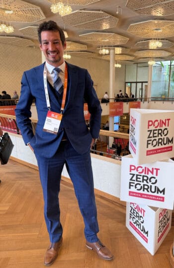 Ein Teilnehmer des Point Zero Forum in Zürich präsentiert sich in einem blauen Anzug mit einer Konferenz-Badge. Im Hintergrund sind weitere Teilnehmer und die Veranstaltungsumgebung sichtbar, die auf eine aktive Diskussion hinweist.
