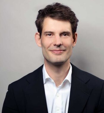 Julius Taylor, Senior Consultant bei Cofinpro, präsentiert sich in einem formellen Outfit. Der Hintergrund ist neutral gehalten. Seine Mimik vermittelt Selbstbewusstsein und Professionalität, was im Kontext der Diskussion über Agentensysteme von Bedeutung ist.