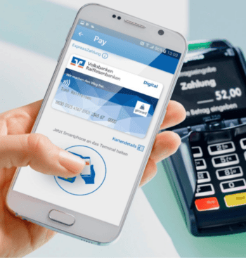 Eine Hand hält ein Smartphone, auf dem eine digitale girocard der Volksbanken Raiffeisenbanken angezeigt wird. Im Hintergrund ist ein Zahlungsterminal zu sehen, das einen Betrag von 52,00 Euro anzeigt. Atruvia spielt eine zentrale Rolle im neuen Kartengeschäft.