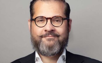 Ein Mann mit Brille und Bart präsentiert sich in formeller Kleidung. Sein Gesichtsausdruck ist freundlich und selbstbewusst. In der Diskussion um Agentensysteme spielt er eine Rolle, indem er effiziente Prozesse in Banken unterstützt.