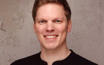 Ein lächelnder Mann mit kurzen, hellen Haaren trägt ein schwarzes Oberteil. Der Hintergrund ist neutral und strukturiert. Die Abbildung könnte im Kontext von Finanzdienstleistern stehen, die sich mit KI-Strategien auseinandersetzen.