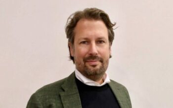 Manuel Sandhoder, ein Vertreter von Wise, ist in einem formellen Outfit abgebildet. Der Hintergrund ist neutral gehalten, was den Fokus auf seine Person lenkt. Seine Mimik vermittelt Professionalität und Engagement im Bereich der Banking-Plattformen.