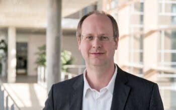 Dr. Christian Schunck, Senior Scientist am Fraunhofer Institut für Arbeitswirtschaft und Organisation IAO, steht in einem modernen Gebäude. Die helle Umgebung und die Pflanzen im Hintergrund unterstreichen eine professionelle Atmosphäre.