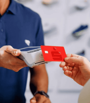 Eine Person reicht eine rote Sparkassen-Card an einen Kassierer, der ein Kartenlesegerät hält. Im Hintergrund sind unscharfe Schuhe zu erkennen. Die Szene deutet auf einen Zahlungsvorgang im Einzelhandel hin.