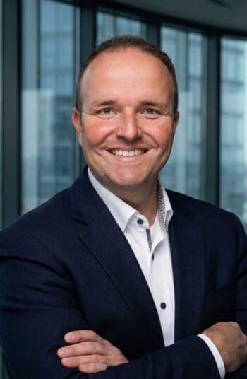 Ein lächelnder Mann mit kurzen Haaren trägt einen dunklen Anzug und ein weißes Hemd. Er steht vor großen Fenstern, die einen Blick auf moderne Gebäude ermöglichen. Der Ausdruck vermittelt Zuversicht und Professionalität, passend zu Finanzprozessen.