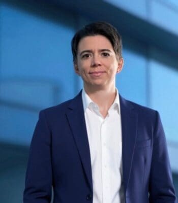 Andrea Woll, neue Leiterin des Schadenmanagements bei die Bayerische, präsentiert sich in einem professionellen Umfeld. Sie trägt einen blauen Blazer und ein weißes Hemd, während der Hintergrund eine moderne Architektur zeigt.