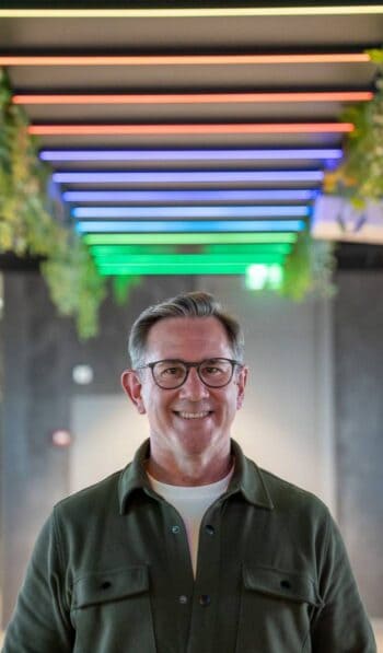 Joe Wilson, Chief Evangelist und ehemaliges Vorstandsmitglied & COO von bunq, steht vor einer modernen, farblich beleuchteten Decke. Diese visuelle Gestaltung spiegelt die Innovationskraft im Bereich BaaS wider und unterstreicht die Rolle von Fintechs im Finanzse