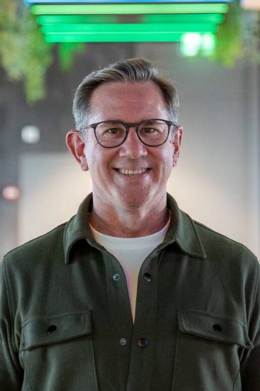 Ein Mann mit Brille und einem freundlichen Lächeln steht in einem modernen Raum. Er trägt ein grünes Oberteil und hat graue Haare. Der Hintergrund ist hell und zeigt eine zeitgemäße Gestaltung, passend zur bunq-Ästhetik.
