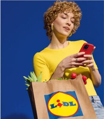Eine Person mit lockigem Haar trägt ein gelbes Oberteil und hält ein Smartphone in der Hand. In der anderen Hand hält sie eine Einkaufstasche mit dem Lidl-Logo, die frisches Gemüse und Obst enthält. Die Integration der Bezahllösung Wero wird hervorgehoben.