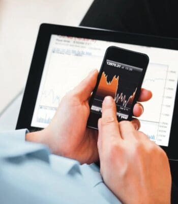 Eine Person nutzt ein Smartphone zur Analyse von Wertpapierkursen, während auf einem Tablet zusätzliche Finanzdaten angezeigt werden. Die App „S-Neo“ ermöglicht den Zugriff auf aktuelle Marktinformationen und unterstützt den Wertpapierhandel.