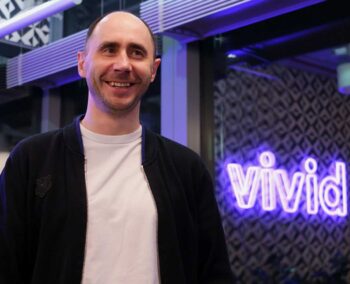 Vivid-Mitgründer Alexander Emeshev steht lächelnd vor einem beleuchteten Vivid-Schild. Die Umgebung ist modern gestaltet, was die Innovationskraft der Berliner Neobank unterstreicht. KI wird hier als integraler Bestandteil des Arbeitsalltags betrachtet.