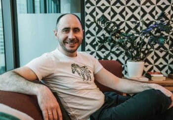 Vivid-Mitgründer Alexander Emeshev sitzt entspannt auf einem Sofa. Er trägt ein T-Shirt mit Zebramuster. Im Hintergrund sind große Fenster und eine Pflanze in einem Topf sichtbar. Die Umgebung wirkt modern und hell.