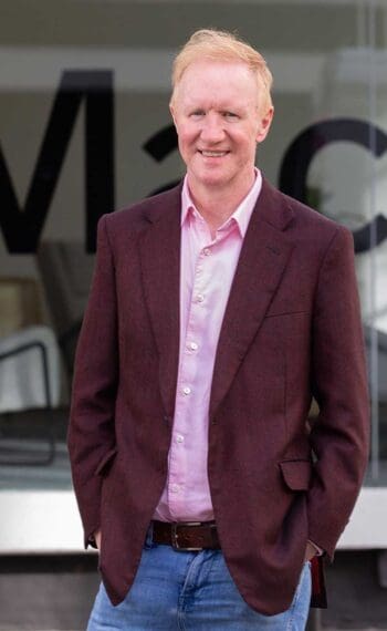 Paul Taylor, CEO und Gründer von Thought Machine, steht in einem modernen Umfeld. Er trägt einen burgunderfarbenen Blazer und ein helles Hemd. Die Umgebung spiegelt eine zeitgemäße Geschäftsatmosphäre wider, die Innovation und Fortschritt symbolisiert.