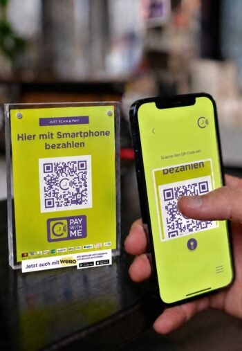 Ein QR-Code auf einem Schild weist darauf hin, dass Zahlungen mit dem Smartphone möglich sind. Neben dem Schild wird ein Smartphone gezeigt, das den QR-Code scannt. Die Lösung „Pay with ME“ von ZIIB ermöglicht Zahlungen mit Wero.