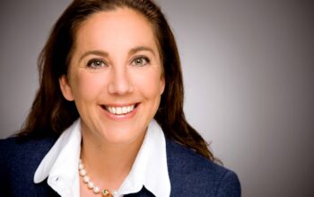 Autorin Dunja Koelwel, IT Finanzmagazin, präsentiert sich mit einem freundlichen Lächeln. Sie trägt eine elegante Bluse und eine Perlenkette. Der Hintergrund ist neutral gehalten, was den Fokus auf ihr Gesicht lenkt.