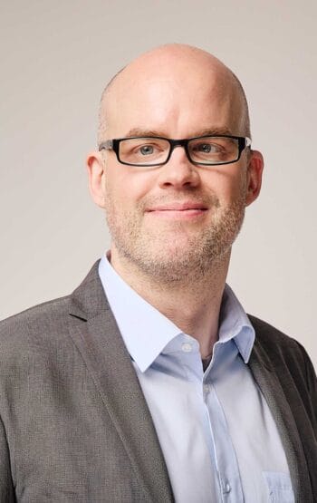 Ein Mann mit Glatze und Brille trägt einen grauen Anzug und ein hellblaues Hemd. Sein Gesichtsausdruck ist freundlich. Im Kontext der digitalen Währungsentwicklung reflektiert er die Vorbereitungen der Banken auf neue Systeme für den Digitalen Euro.