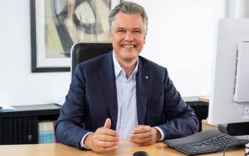Dr. Herbert Schneidemann, Vorsitzender des Vorstands der Bayerischen, sitzt an einem Schreibtisch und lächelt. Im Hintergrund sind abstrakte Kunstwerke und Büromöbel sichtbar. Ein Computer und eine Tastatur sind auf dem Tisch platziert.
