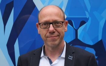 Michael Brand, ein IT-Experte mit langjähriger Erfahrung in der Versicherungswirtschaft, steht vor einer blauen, geometrischen Wand. Seine Expertise in Software-Engineering ist relevant für die Diskussion über Zero Trust und die Notwendigkeit von PAM.