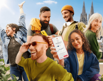 Eine Gruppe junger Menschen interagiert fröhlich im Freien, während sie Smartphones nutzen. Im Vordergrund hält eine Person ein Mobilgerät mit einer Finanzanwendung der Sparkasse KölnBonn. Im Hintergrund sind die Kölner Domtürme sichtbar.