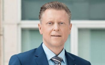 Stefan Schulz, AML-Officer der BarmeniaGothaer, präsentiert sich in einem formellen Anzug. Der Hintergrund zeigt eine moderne Architektur, die Professionalität und Seriosität ausstrahlt. Sein Blick ist direkt und selbstbewusst, was seine Rolle unterstreicht.