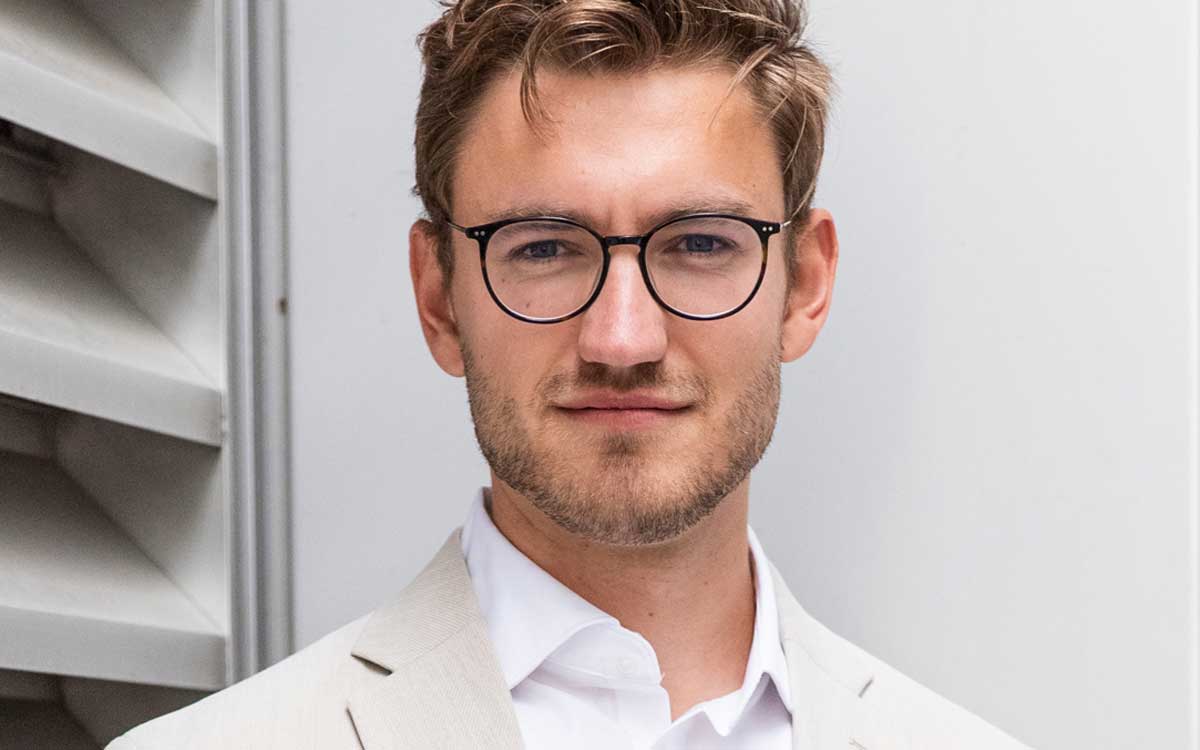 Tobias Weitzel, Head of confora labs bei spotixx, präsentiert eine Anleitung zur Fairness in Hochrisiko-KI. Sein professionelles Auftreten wird durch einen hellen Anzug und eine Brille unterstrichen, vor einer neutralen Hintergrundstruktur.