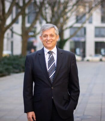 Anastasios Papakostas, neuer Vice President of Business Development bei enclaive, steht in einem urbanen Umfeld. Er trägt einen dunklen Anzug mit gestreifter Krawatte und lächelt, während er entspannt mit einer Hand in der Hosentasche posiert.