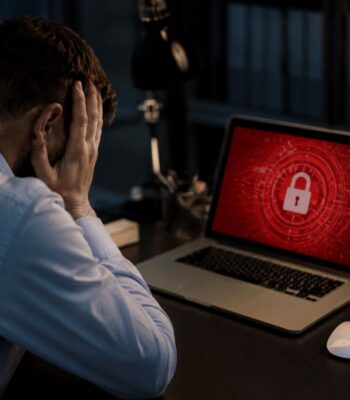 Ein Mann mit einem Hemd sitzt an einem Schreibtisch und hält sich den Kopf in den Händen. Auf dem Laptop-Bildschirm ist ein rotes Hintergrundbild mit einem Schloss-Symbol zu sehen, was auf Cyberrisiken hinweist.