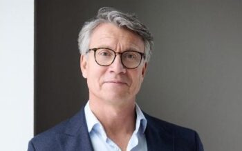 Andreas Gerhardt steht im Porträt vor einer grauen Wand neben einem hellen Fenster. Er trägt Brille, dunkelblaues Sakko und hellblaues Hemd und blickt freundlich in die Kamera.