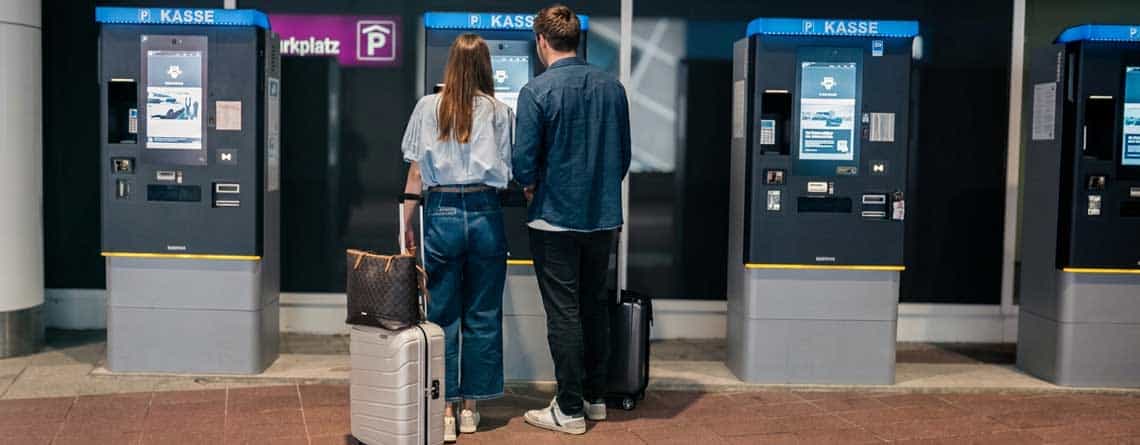 Zwei Reisende stehen vor Parkautomaten am Flughafen München. Die Automaten bieten verschiedene Zahlungsoptionen, darunter die Integration von Epay. Im Hintergrund sind Hinweise auf den Parkplatz sichtbar.