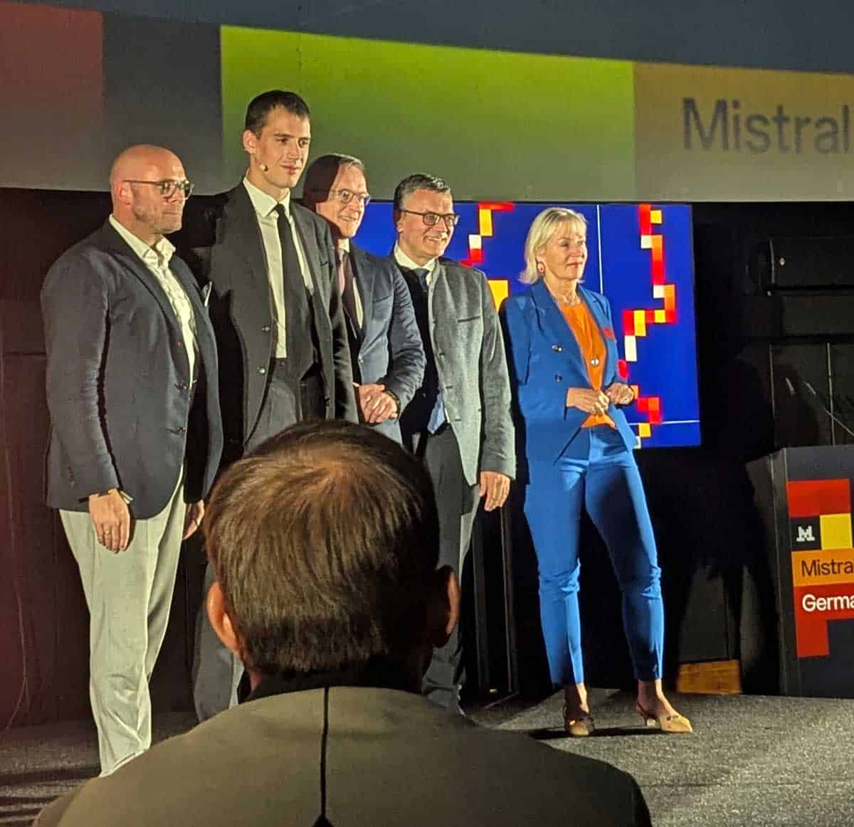 Auf der Veranstaltung in München präsentieren sich mehrere Personen, die mit Mistral in Verbindung stehen. Im Hintergrund ist ein Bildschirm mit dem Logo von Mistral sichtbar. Die Atmosphäre ist professionell und feierlich.