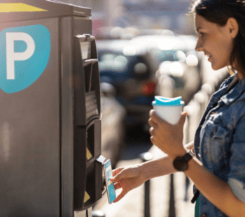 Eine Person nutzt einen Parkautomaten, um bargeldlos zu bezahlen. In der Hand hält sie einen Becher und eine girocard wird zur Zahlung verwendet. Im Hintergrund sind parkende Fahrzeuge sichtbar.