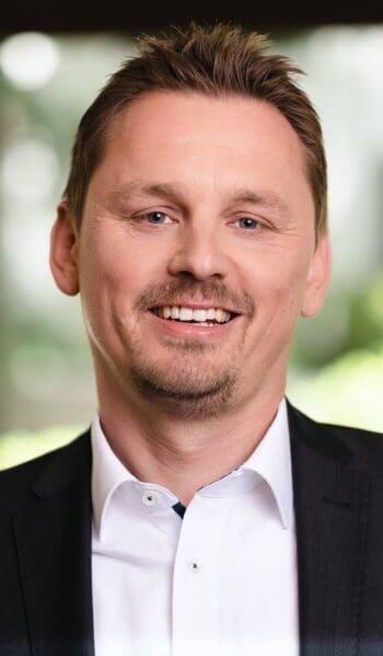 Christian Koch, Senior Vice President Cybersecurity IoT/OT, Innovations und Business Development bei NTT DATA DACH, präsentiert sich in einem professionellen Umfeld. Sein Lächeln und die formelle Kleidung unterstreichen seine Rolle in der Banken-IT und den Herausforderungen im Bereich Cybersecurity.