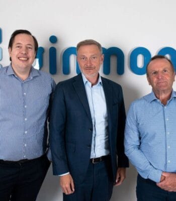Christian Lindner, Bundesminister der Finanzen, steht zwischen zwei weiteren Personen vor dem Logo von Ginmon. Die Anwesenden tragen formelle Kleidung und lächeln, was auf eine positive Atmosphäre hindeutet.