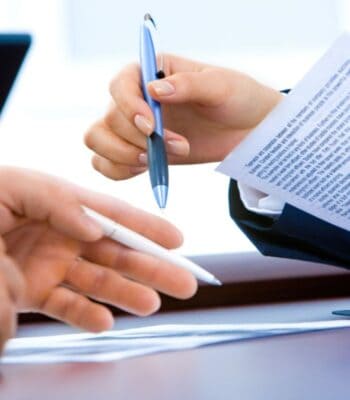 Zwei Hände sind in einem geschäftlichen Kontext abgebildet. Eine Hand hält einen blauen Stift, während die andere Hand einen weißen Stift hält. Im Hintergrund sind Dokumente sichtbar, die möglicherweise mit fino digital in Verbindung stehen.