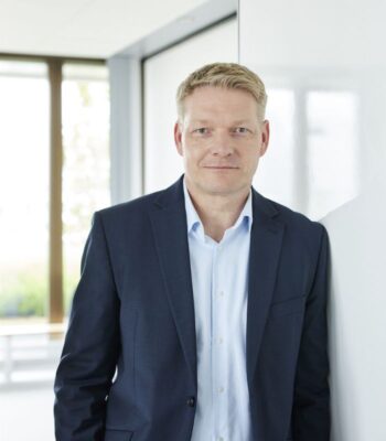 Stefan Roesler, neuer Geschäftsführer bei Finanz Informatik, steht in einem modernen Büro. Er trägt einen dunkelblauen Anzug und blickt direkt in die Kamera. Die Umgebung ist hell und freundlich, mit großen Fenstern im Hintergrund.