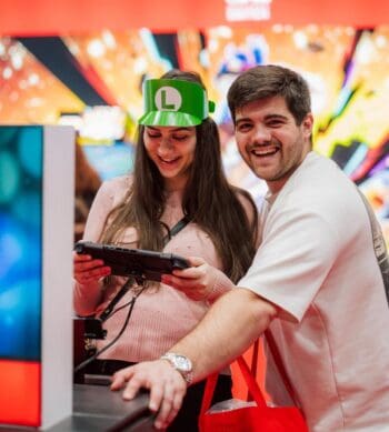 Eine Frau mit grüner L-Kappe spielt an einer Konsole, während ein Mann lächelnd danebensteht; auf einer Gaming-Messe wird gezockt. Gaming erreicht Millionen Menschen.