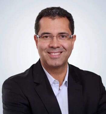 GFT-CEO Marco Santos mit Brille, dunklem Sakko und hellem Hemd vor hellem Hintergrund; er spricht über KI-gestützte Batch-Migration vom Mainframe in die Cloud.