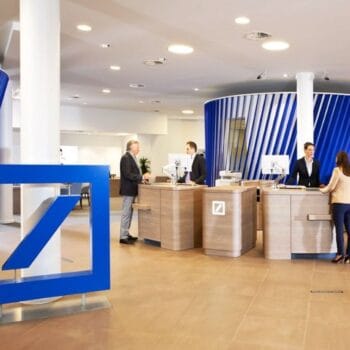 Kunden sprechen mit Bankmitarbeitenden an Schaltern in einer Deutsche-Bank-Filiale; die Bank erweitert ihre Vermögensverwaltung um einen digitalen Abschluss über die Banking-App.