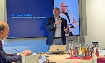 Digitalminister Dr. Fabian Mehring präsentiert während einer Veranstaltung in München. Im Hintergrund ist eine Präsentation zu sehen. Auf dem Tisch stehen verschiedene Getränke, während ein Teilnehmer aufmerksam zuhört.
