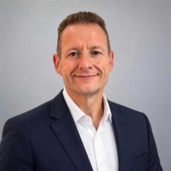 Graham Clarke, Managing Director Global Private & Sovereign AI bei NTT Data, präsentiert sich in einem professionellen Umfeld. Sein Lächeln und die formelle Kleidung unterstreichen die Bedeutung der Eröffnung der AI Factory in München.