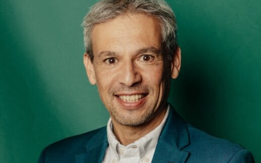 Hans Jayatissa lächelt im weißen Hemd und türkisfarbenen Sakko vor grünem Hintergrund; als Quantum Technology Expert befasst er sich mit Quantensicherheit in der Finanzbranche.