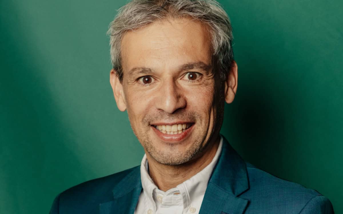 Hans Jayatissa lächelt im weißen Hemd und türkisfarbenen Sakko vor grünem Hintergrund; als Quantum Technology Expert befasst er sich mit Quantensicherheit in der Finanzbranche.