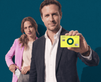 Eine Person präsentiert eine gelbe Holvi-Karte, während eine weitere Person im Hintergrund steht. Der Hintergrund ist einfarbig in Blau gehalten. Die Darstellung vermittelt den Fokus auf die angebotenen Finanzdienstleistungen von Holvi.