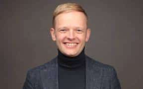 Jan Wichmann sitzt lächelnd vor grauem Hintergrund, in dunklem Rollkragenpullover und kariertem Sakko. Als Gründer von JWR Advisory berät er zum BaaS-Ausstieg und zu Nachfolgelösungen für IT-Dienstleister.