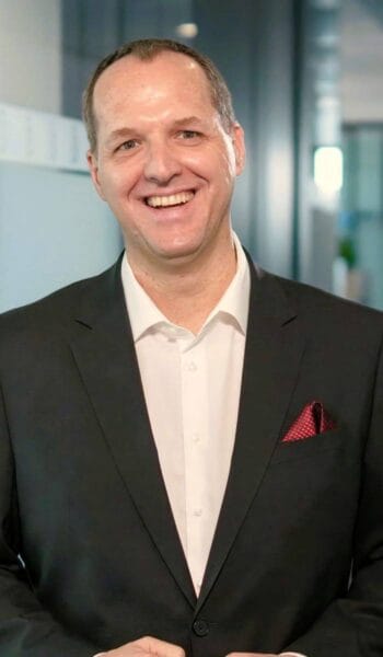 Tom Würfel, Senior Director Solution Sales DACH bei Dun & Bradstreet, lächelt im dunklen Anzug vor einer Glaswand; er erläutert, wie KI-Agenten in Bank- und Versicherungs-IT zusammenspielen.