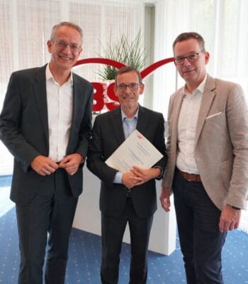 Drei Personen stehen vor einem Hintergrund mit dem Logo der LBS. Eine Person hält ein Dokument in den Händen. Die Atmosphäre wirkt formal und geschäftlich, was auf die Verlängerung der Partnerschaft zwischen LBS NordWest und LBS Hessen-Thüringen hinweist.